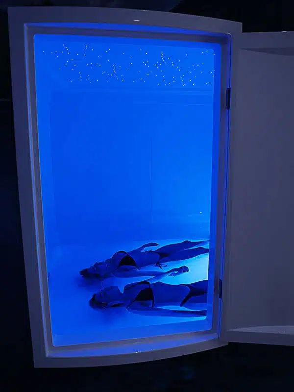 Couple holding hands floating in a double Ocean Float Room surrounded in soft blue light